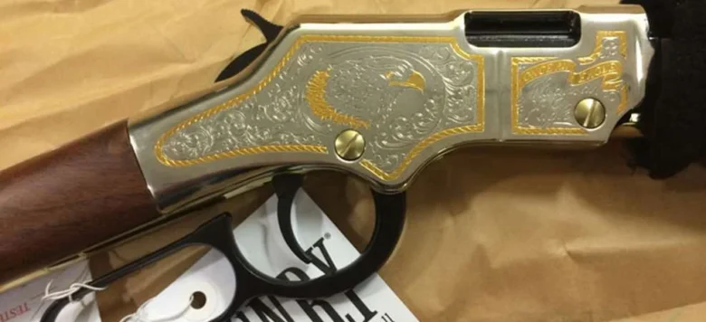 Decorative lever-action firearm with silver and gold engraving, wooden stock, lying on a beige surface with a Henry tag attached.