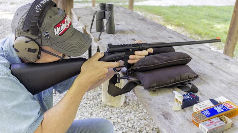Man wearing a cap and ear protection aims a bolt-action rifle on a wooden shooting bench, with ammo boxes on the table nearby.