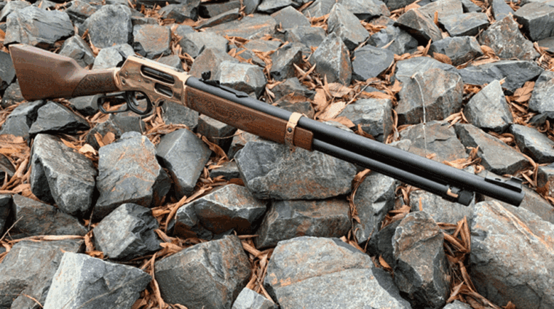 Shotgun with wooden stock and long barrel resting on gray rocks on a rocky ground surface.