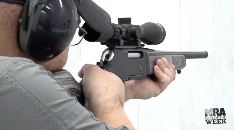 Shooter aiming a scoped rifle, wearing ear protection, at a target.