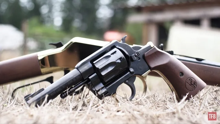 A black revolver resting on dry grass outdoors, with a rifle partially visible in the background.