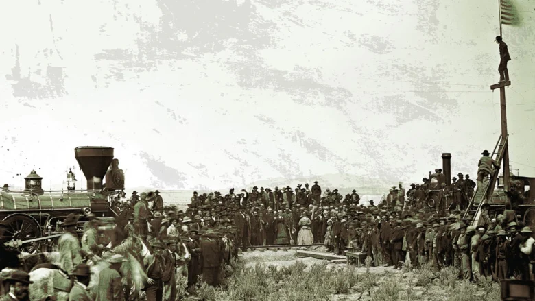 Historic crowd gathered near a steam locomotive for a ceremony, with a man on a tall platform ladder above the onlookers.