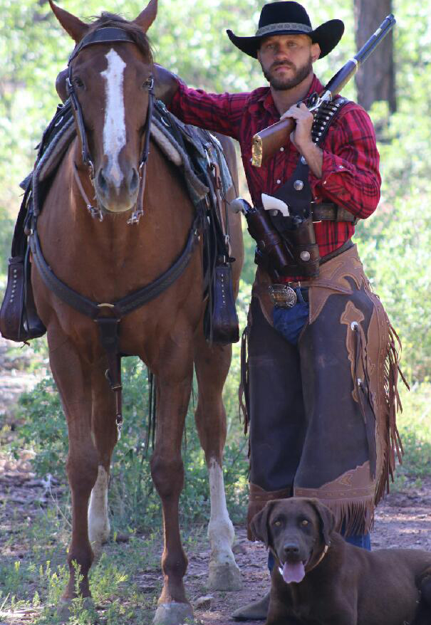 Cowboy with rifle standing next to horse and hunting dog in outdoor setting