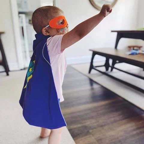 Young child dressed in superhero costume, arms raised, playing in living room
